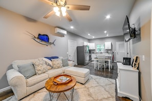 A modern living room with a sofa, coffee table, and a ceiling fan. There is a kitchen area with cabinets and a refrigerator in the background.