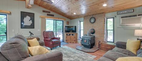A cozy cabin interior with wooden ceilings, a stone wall, a fireplace, and a large window.