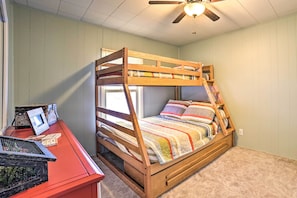 Un lit superposé avec une structure en bois et une literie colorée, un bureau avec un ordinateur et des livres, et un ventilateur de plafond.