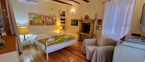 A cozy bedroom with a wooden floor, a bed with white bedding, a small table with a lamp, a chair, and a fireplace.