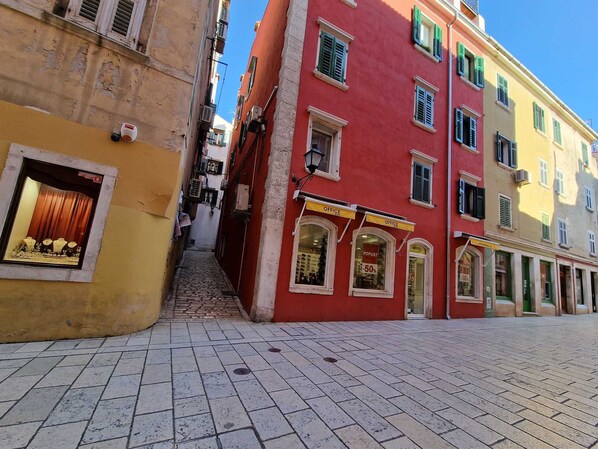 A narrow street with colorful buildings, a shop with a sign 'POPUST - 50% OFF', and a small staircase leading to a door.