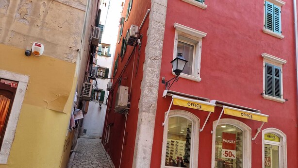 Una stretta strada acciottolata con un edificio rosso che ospita un negozio 'Office Shoes' e un cartello che indica uno sconto del 50%.