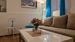 A living room with a white sofa, a glass coffee table, a metal chair, a vase with flowers, a painting on the wall, and a floor lamp. - LIMONA - Studio Apartment (Rovinj)