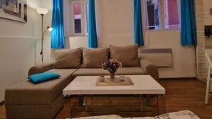 A living room with a beige sofa, a white coffee table, and a vase with flowers. There are blue curtains on the windows, and a floor lamp is visible. - LIMONA - Studio Apartment (Rovinj)