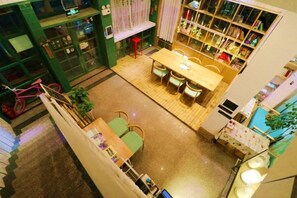 A dining area with a wooden table and chairs, a staircase, and a bookshelf with various items. - Xiyu Hostel · Cat Café (Yangshuo)