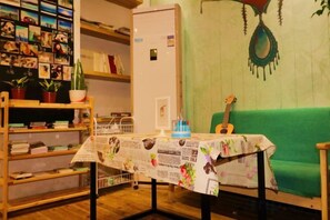 A room with a green couch, a guitar, a table with a newspaper tablecloth, and a bookshelf filled with books and pictures. - Xiyu Hostel · Cat Café (Yangshuo)