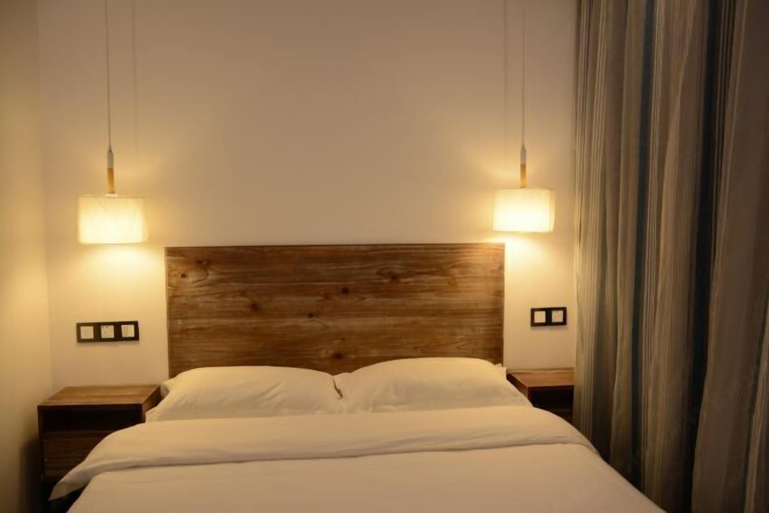 A hotel room with a wooden headboard, two bedside lamps, a bed with white bedding, and a curtain on the right side.