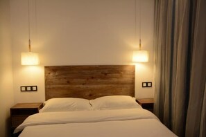 Habitación de hotel con cabecera de madera, dos lámparas de noche, cama con ropa blanca y una cortina a la derecha.