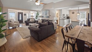 A spacious living area with a dark grey sofa, a wooden dining table, and a kitchen in the background.