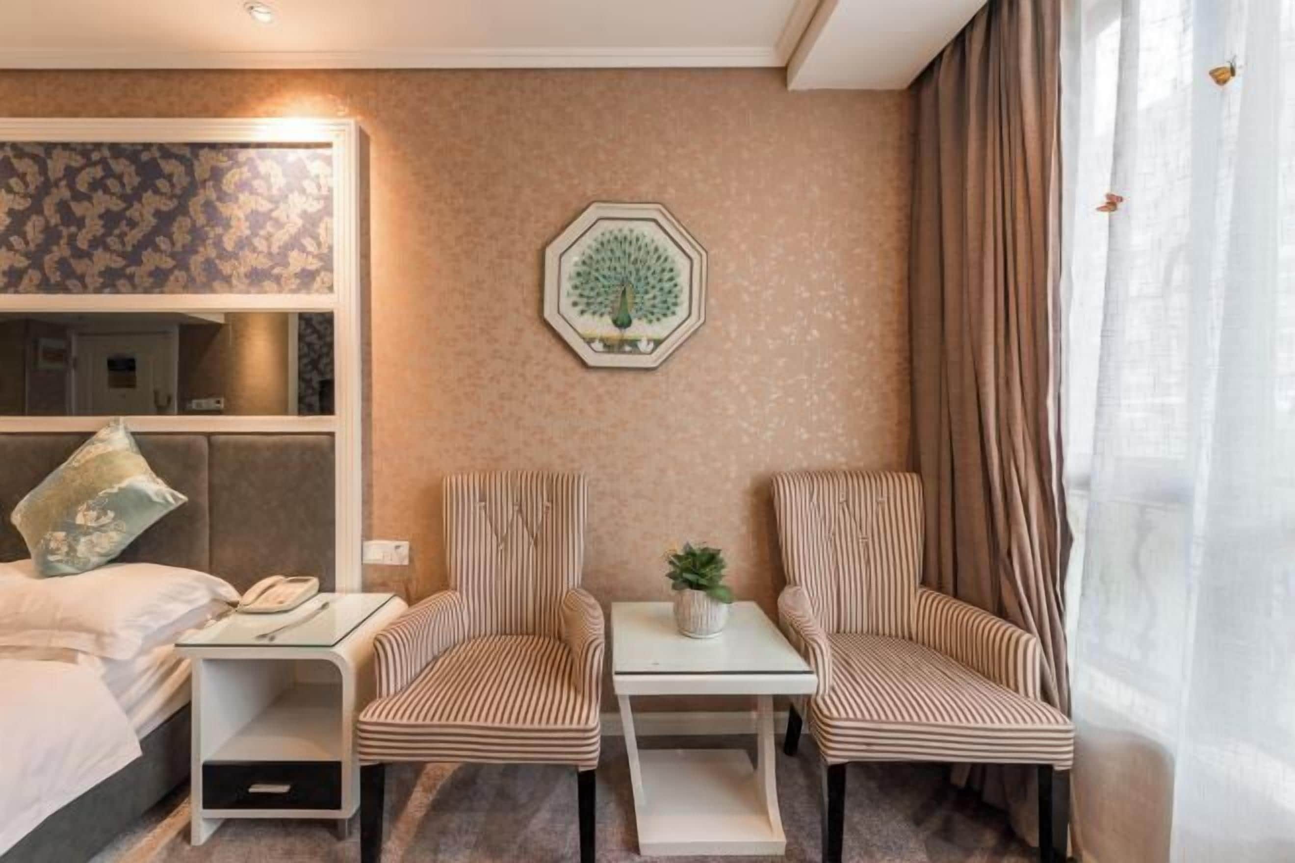 Une chambre d’hôtel avec un lit, deux fauteuils à rayures, une petite table avec une plante, un miroir et un élément décoratif mural.