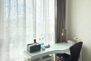 Habitación de hotel con escritorio de vidrio, una silla negra, teléfono, lámpara y ventana con cortinas translúcidas.