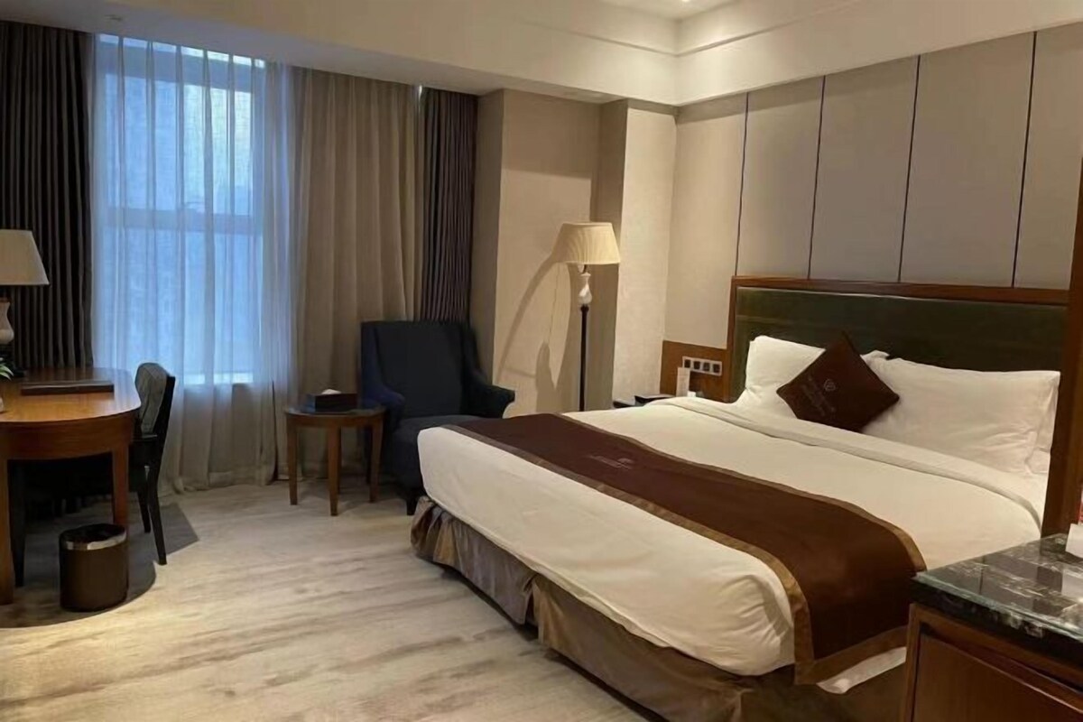A hotel room with a large bed, a desk with a chair, a small table, and a lamp.