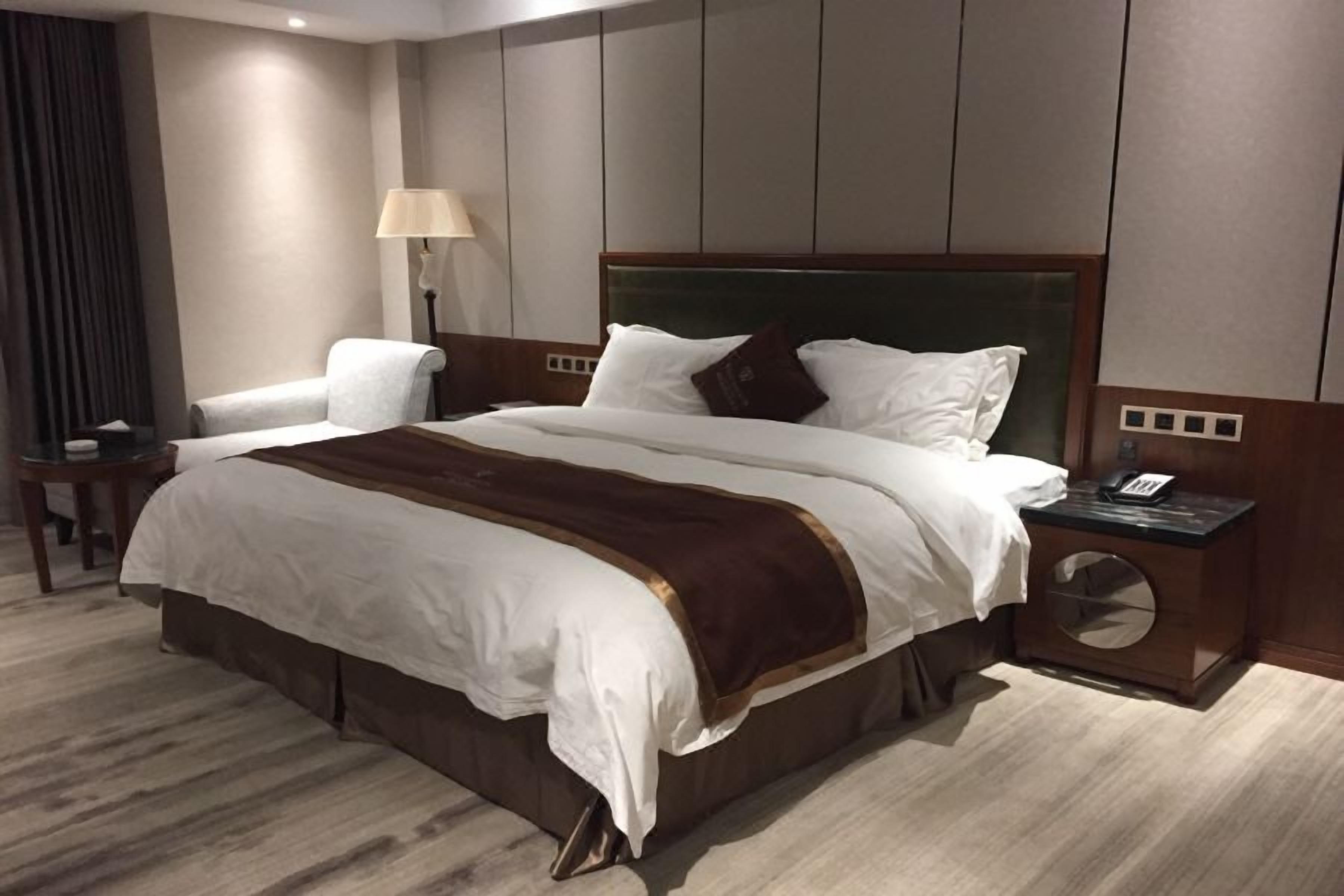 A hotel room with a large bed, a bedside table with a lamp, a chair, and a round mirror on the nightstand.