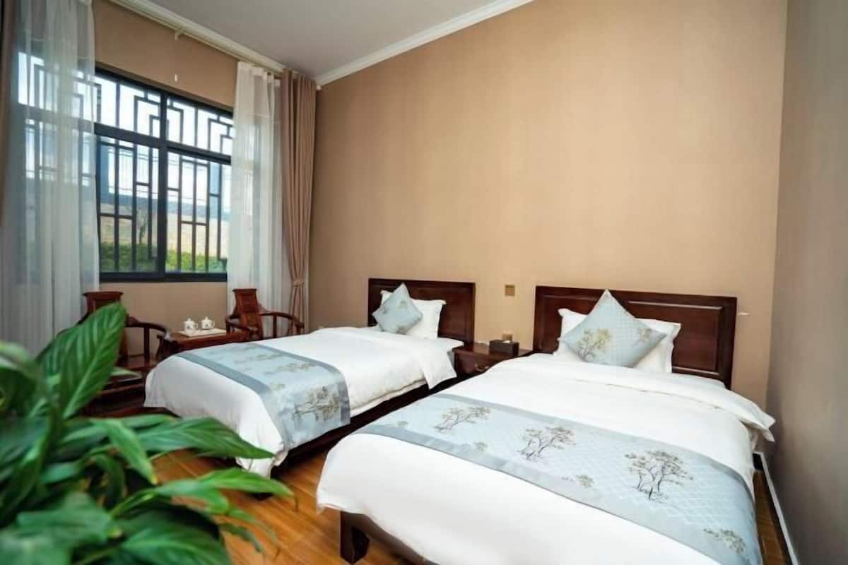A hotel room with two beds, a window with curtains, a small table with a teapot, and a potted plant.