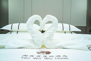 A bed with white linens and a heart-shaped towel arrangement. The bed has a white pillow with a brown stripe and a white pillow with a brown stripe. The text on the bed reads 'Tao Wo Wo Hotel.' - Taowowo Holiday Hotel (Jianshui)