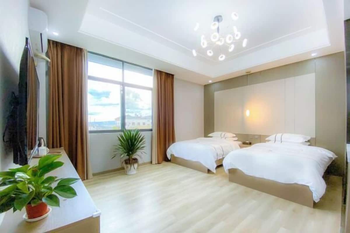 A hotel room with two beds, a large window, a ceiling light fixture, and a potted plant.