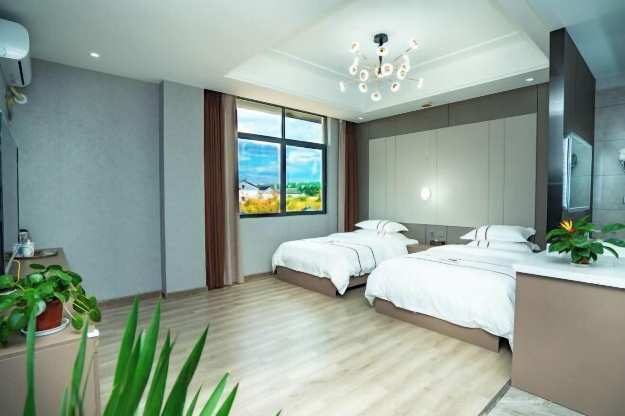 A modern hotel room with two beds, a large window, a ceiling light, and a plant.