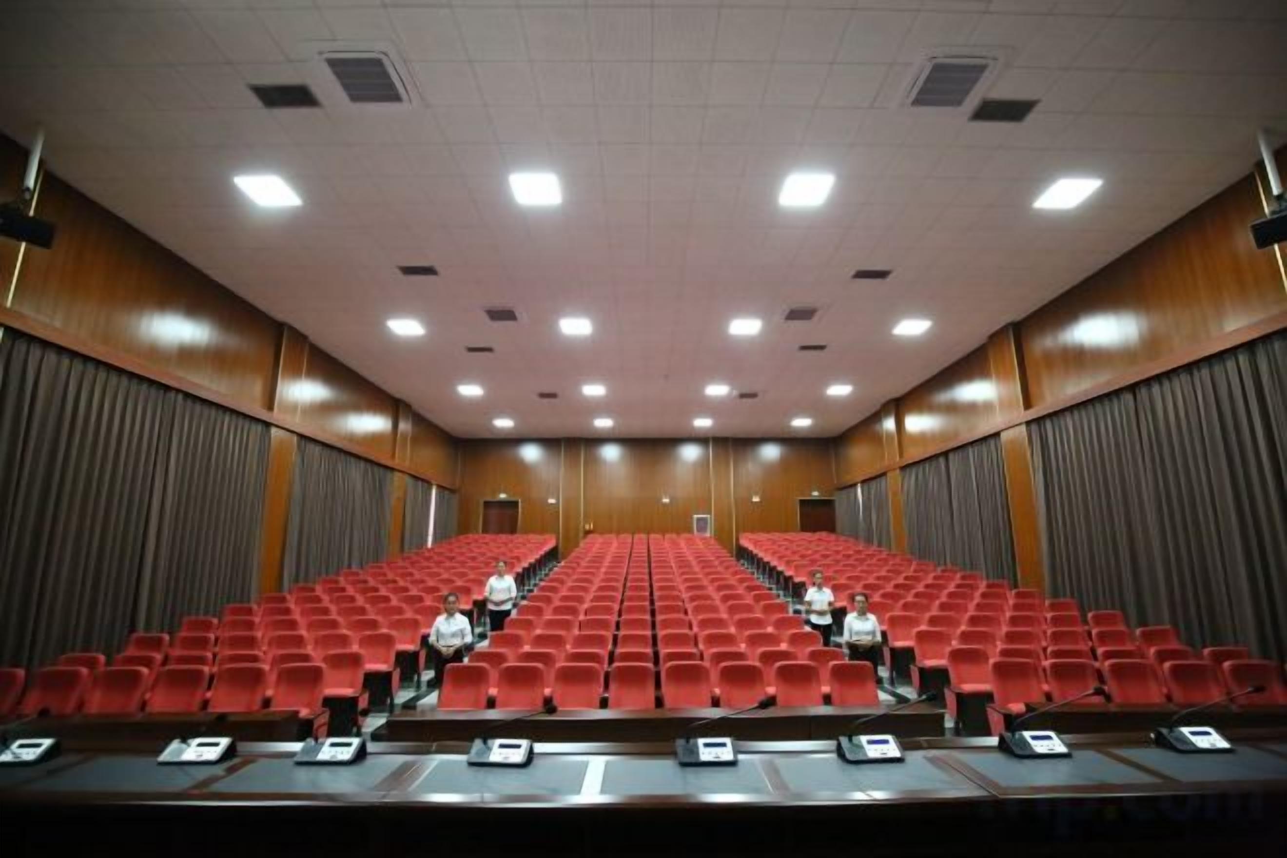 Un salón de conferencias con filas de sillas rojas, un atril al frente y una pantalla grande.