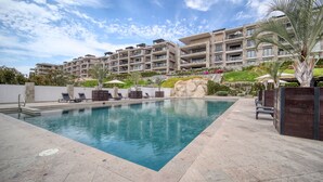 Una piscina con sillones de descanso, un muro de piedra y un edificio de varios pisos con balcones al fondo.