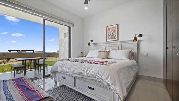 A bedroom with a large bed, a nightstand, a wall-mounted lamp, and a view of the outdoors.