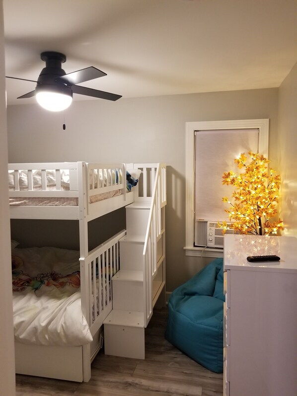Una habitación con una cama individual superpuesta, un ventilador de techo, una mesita con una planta decorativa y un sillón de pelota azul.
