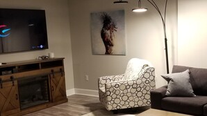 Una sala de estar con televisión, un sillón estampado, un sofá, una mesa de centro, chimenea y un cuadro en la pared.