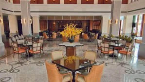 Un lobby espacioso con una mesa redonda central rodeada de sillas de mimbre, un jarrón grande con flores amarillas y un piso con diseño.