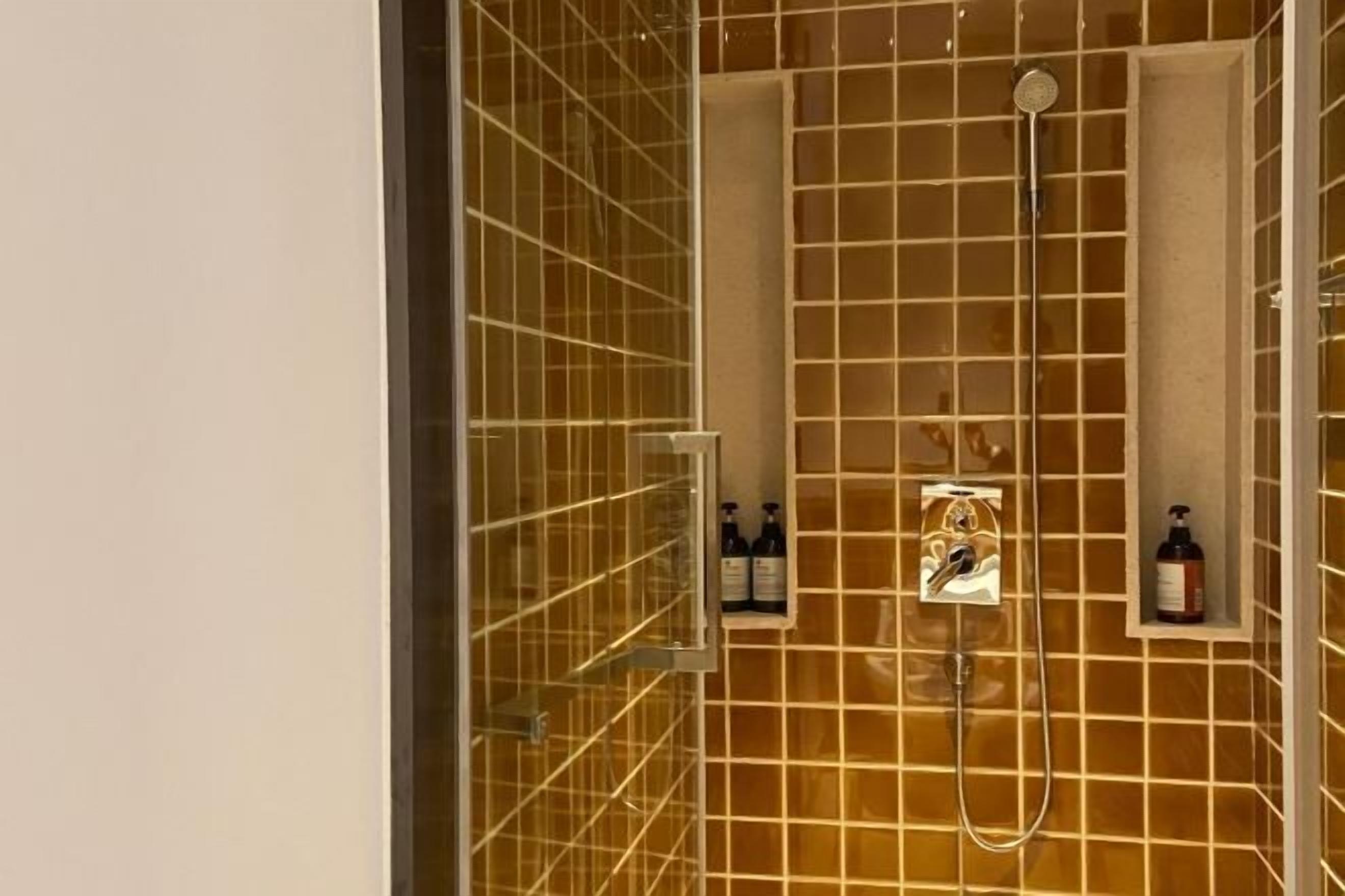 Ducha con puerta de vidrio, cabezal de ducha manual y dos botellas de champú en la pared.