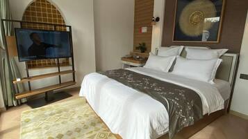 Habitación de hotel con cama, televisor sobre un mueble, una mesita con una planta y una obra de arte colgada en la pared.
