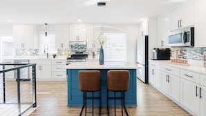 Una cocina moderna con armarios blancos, una isla azul, dos taburetes de bar y un refrigerador de acero inoxidable.