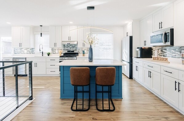 Une cuisine moderne avec des armoires blanches, un îlot de couleur bleue, deux tabourets de bar et un réfrigérateur en acier inoxydable.