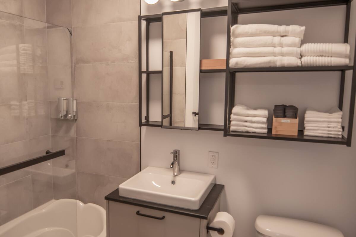 A modern bathroom with a glass-enclosed shower, a white sink with a black countertop, and a toilet. There are shelves with neatly folded white towels and a mirror above the sink.