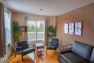 Una sala de estar moderna con un sofá, dos sillones, una mesita de centro y un ventanal con cortinas. Hay dos plantas en macetas y un estante en la pared con objetos decorativos.