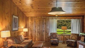 A cozy room with wooden walls and ceiling, featuring a sofa, two armchairs, a wooden coffee table, and a desk with a lamp. A window with curtains offers a view of greenery outside. - Two bedroom log cabin (Ennis)