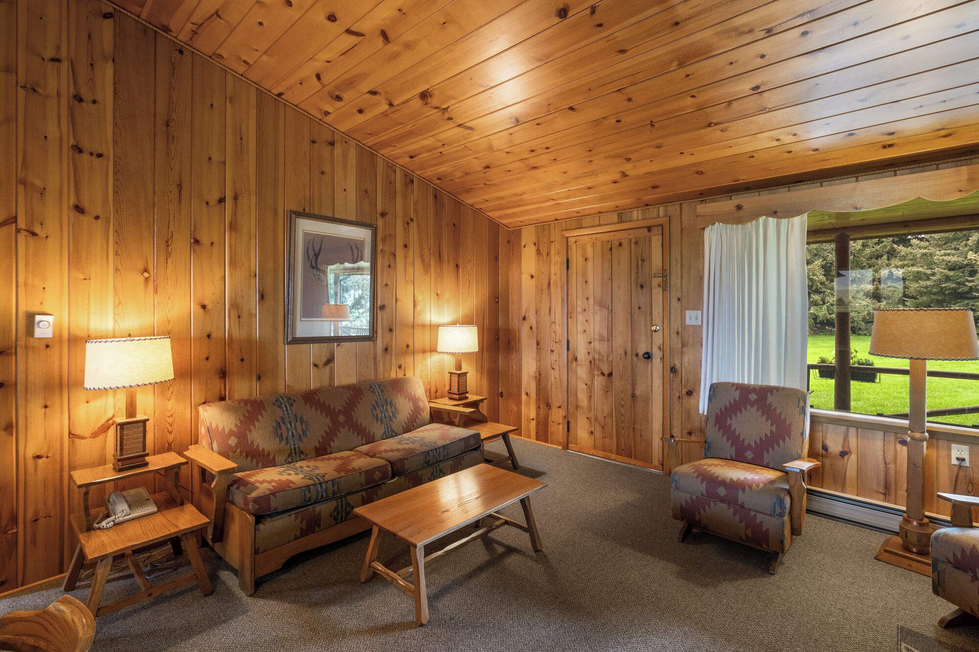 A cozy cabin interior with wooden walls and ceiling, a sofa, a chair, a small table, and a window with curtains.
