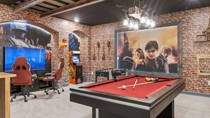 Ein Billardtisch mit roter Spielbahn, ein Kicker, ein PS5 Gaming-Sessel, ein großer Flachbildfernseher und ein Filmplakat an der Wand.