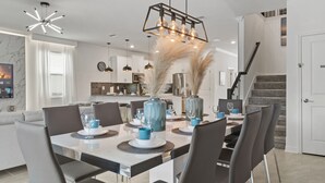 A modern dining area with a white table, dark chairs, and blue accents. There is a kitchen in the background with white cabinets and a stainless steel refrigerator.