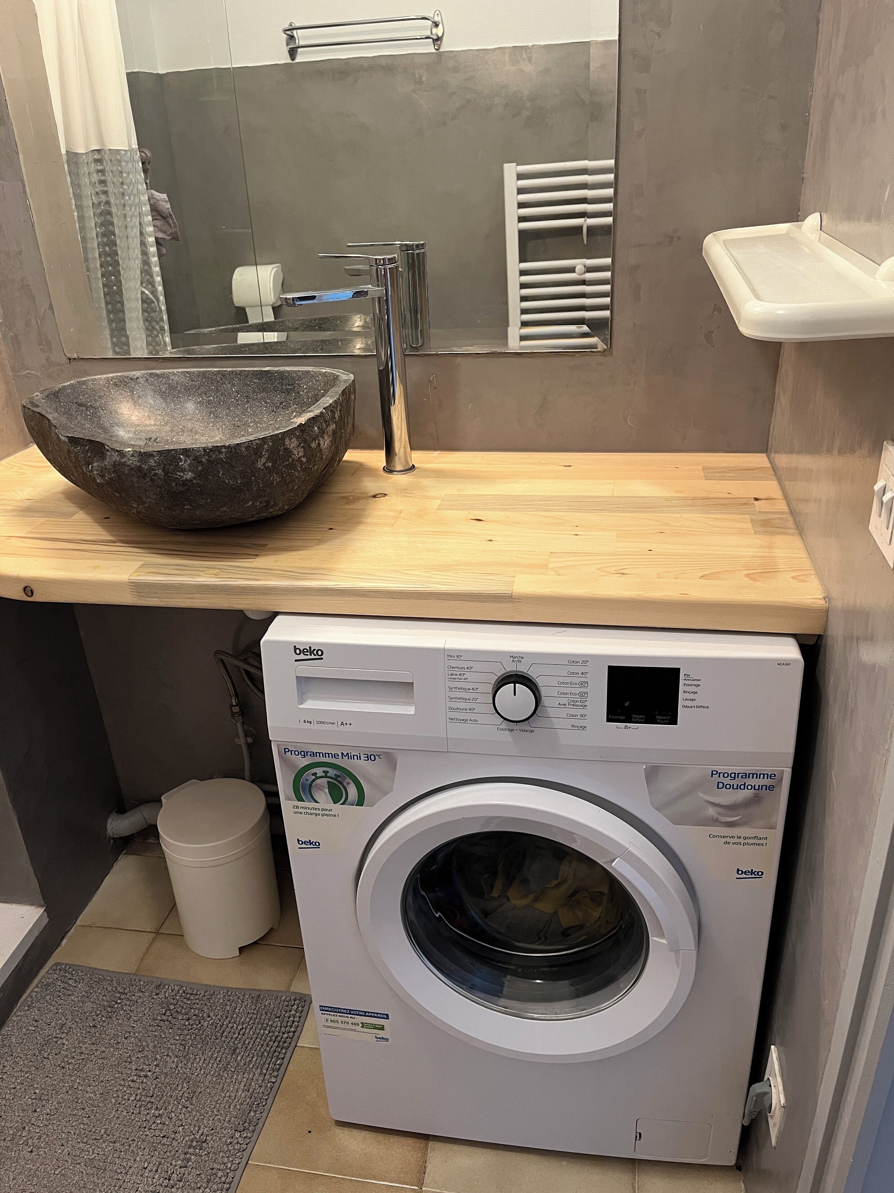 Een kleine badkamer met een stenen wastafel, een moderne kraan, een ingebouwde wasmachine en een kleine prullenbak.