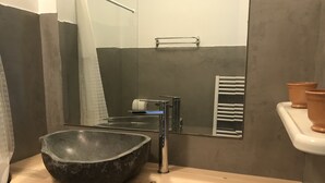 Un baño moderno con un lavamanos de piedra grande, un espejo, un perchero y una pequeña estantería con macetas de terracota.