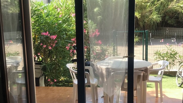 Un patio con mesa y sillas, rodeado de follaje y flores rosadas.