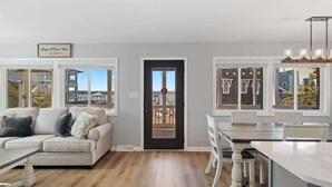 Una sala de estar moderna con un sofá, una mesa de comedor y sillas, una zona de cocina con mostrador, y ventanas amplias que ofrecen vistas al exterior.