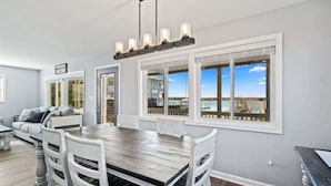 Un comedor con una mesa de madera y sillas blancas, ventanas amplias con vistas, y una lámpara colgante.