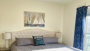 A bedroom with a tufted headboard, a bed with a grey quilt, a pillow with a quote, a painting of sailboats, a ceiling fan, and a window with blue curtains. - Walnut Beach Inn - close to the beach! (Ashtabula)