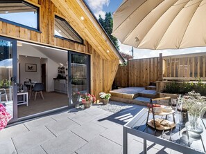 Une maison en bois avec une porte en verre donnant sur un coin repas, une terrasse avec une table et des chaises, et un grand parasol.