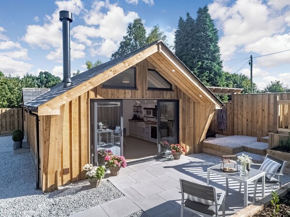 A wooden cabin with a glass door entrance, a patio with seating, and a chimney.