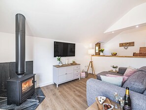 Ein Wohnzimmer mit einem schwarzen Kaminofen, einem grauen Sofa, einem weißen Schrank, einem Wand-Fernseher und einem Holzregal mit diversen Gegenständen.