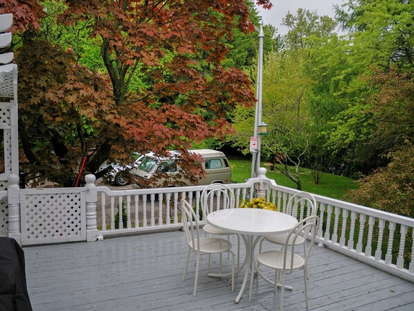 Un porche cubierto con una mesa y sillas blancas, rodeado por una valla blanca, con vistas a un jardín con árboles y un coche estacionado.