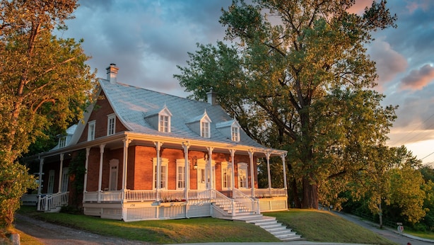 A large, two-story brick house with a wrap-around porch and a chimney. - Appartement Jacques-Cartier (Saguenay)