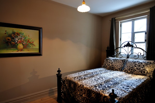 A bedroom with a bed, a window with curtains, a painting on the wall, and a hanging light fixture. - Appartement Jacques-Cartier (Saguenay)