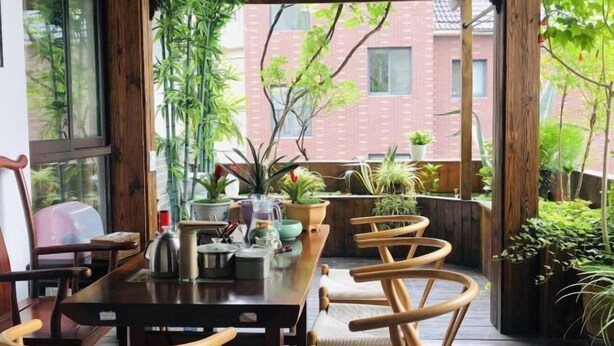 A wooden dining table with chairs, surrounded by potted plants and a view of a brick building. - Pailou Guesthouse (Yueqing)